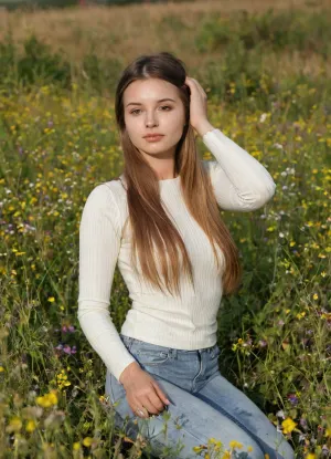 Amature candid photo, ultimate realism, young woman with long, straight brown hair sits in a field of wildflowers, wearing a tight, ribbed, long-sleeve top and high-waisted jeans. She is looking directly at the camera with a neutral expression, holding her hair up with one hand. The lighting is soft and natural, with a warm, golden hue. The background is blurred, showing a field of tall grass and wildflowers. The image has a shallow depth of field, focusing on the woman. The camera angle is slightly low, looking up at her.
