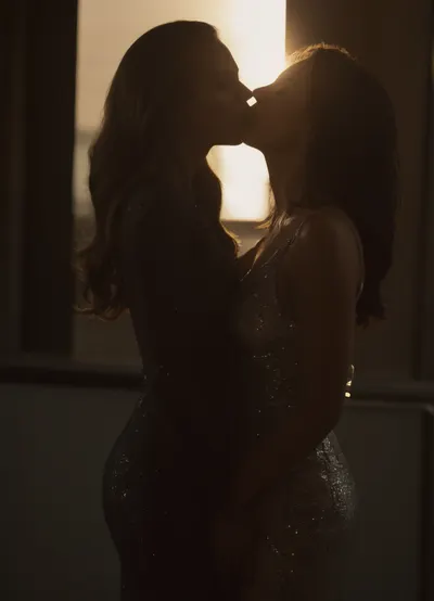Two elegant women in stunning sequined evening gowns—one in deep navy blue, the other in shimmering silver-gray—standing close and facing each other intimately. They passionately kiss, their gazes locked, conveying deep emotion. Each woman gently put a hand under the other's upturned dress and rub the hand against the other's pussy. Both pussies are very wet, pussy juice is flowing down their legs, both aroused, highlighting their slender, graceful figures. Their makeup is flawless and glamorous, emphasizing natural beauty with subtle smoky eyes and soft, sculpted lips. Their hair cascades smoothly, one with long, soft blonde waves illuminated by warm, golden sunlight streaming through a large glass window, creating radiant highlights and delicate glimmers on their sparkling dresses. They each The setting is a cutting-edge modern architectural space featuring sleek geometric shapes, clean lines, and reflective surfaces in neutral, sophisticated tones, enhancing the luxurious and romantic atmosphere. Warm, dynamic lighting emphasizes textures and contours, casting soft sunbeams that play upon their hair and attire, adding a captivating glow and depth to the scene. The overall mood is dramatic, intimate, and artistically refined.